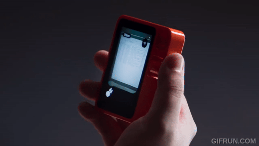 A hand holding an a model of the Rabbit R1 AI assistant device with the screen showing the device performing some action, against a dark background.